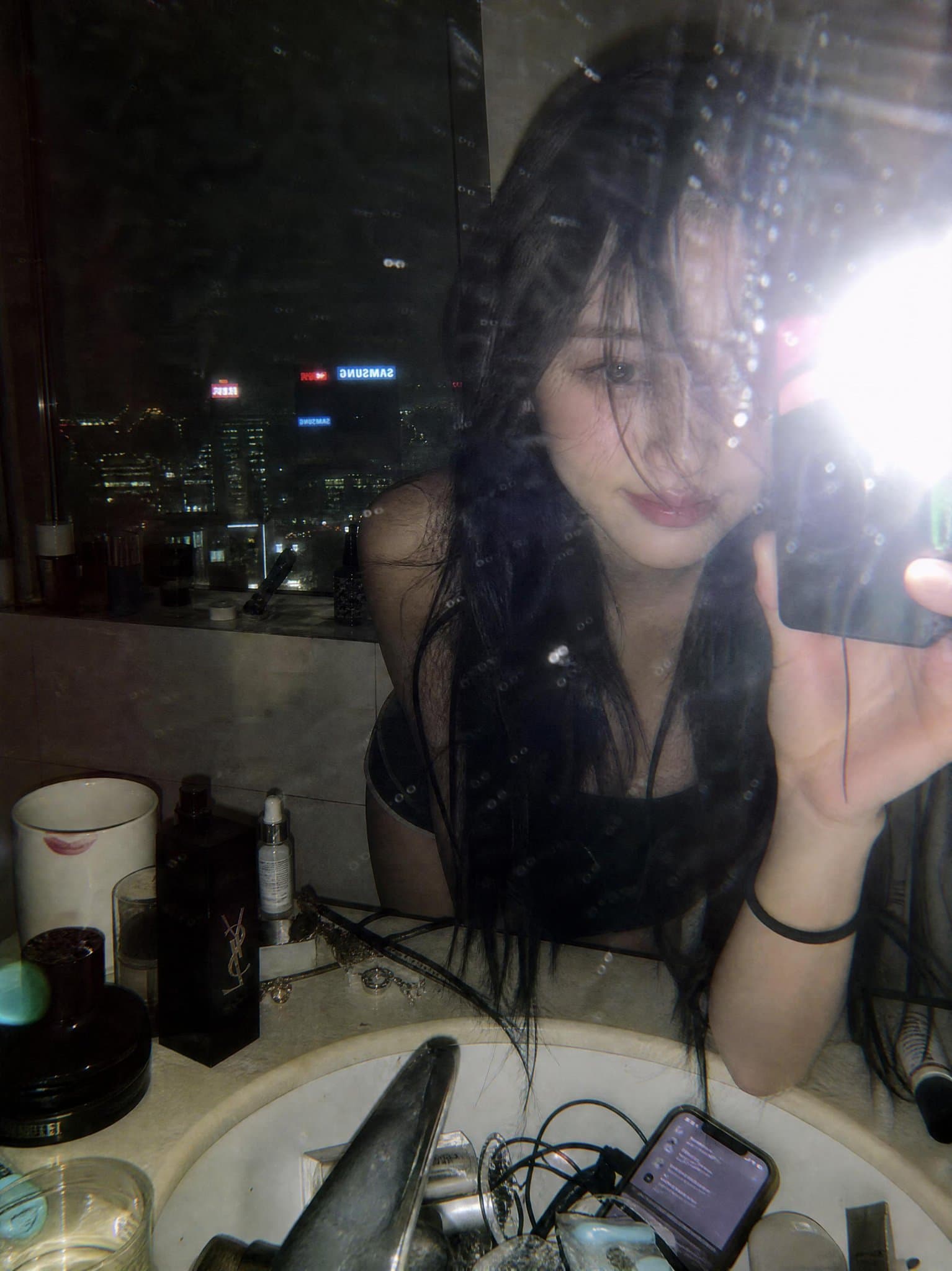 다이렉트 플래시, 얼룩진 유리, 도시 스카이라인 반사, 카운터 위 럭셔리 코스메틱이 있는 Y2K 하이라이즈 욕실 미러 셀피.